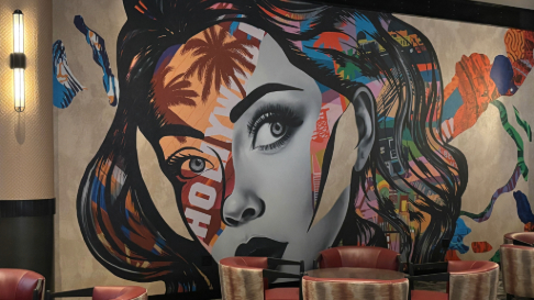 A large, vibrant mural dominates the wall of a modern lounge area, featuring a stylized close-up portrait of a person’s face rendered in a mix of grayscale and bold, multicolored patterns. One eye is highly detailed and realistic, while the rest of the face is overlaid with abstract shapes, palm tree imagery, and bright colors such as orange, blue, red, and yellow that blend into flowing, hair-like forms extending outward. In front of the mural are several low, round tables and red upholstered chairs arranged for seating. The space includes warm lighting and textured wall surfaces, giving the room a contemporary, artistic atmosphere.